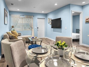 A modern living room with a dining area, light blue walls, a flat-screen TV, and a visible door leading to a bedroom. - Blue Haven Retreat (Sarasota)