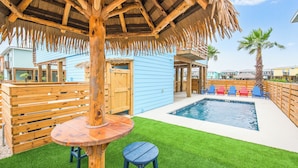 Een zwembadgedeelte met een houten tafel en stoelen, een overkapping met rieten dak en een blauw gebouw op de achtergrond.