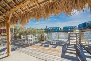 A wooden deck with chairs, a thatched roof, and a view of buildings across a body of water. - Why Knot Relax - Heated Private Pool! Beach Gear Credit Included (Fresno)
