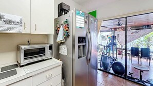 Uma cozinha compacta com micro-ondas, torradeira e frigorífico. Uma pequena janela mostra um pátio com área de estar ao ar livre e uma bicicleta.