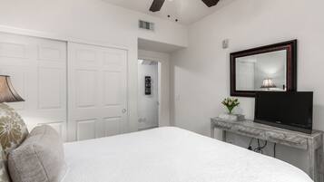 A bedroom with a bed, a TV on a console table, a ceiling fan, and a door leading to another room.