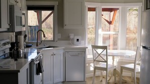 A kitchen with white cabinets, a dishwasher, a kettle, and a dining area with a table and chairs. - Skyriver Escape Riverfront With Hot Tub (Monroe)