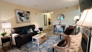 Un salon comprenant un canapé en cuir noir, une table basse, un fauteuil beige, une table à manger avec des chaises et un téléviseur fixé au mur.