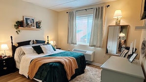 Une chambre à coucher comprenant un lit, des tables de chevet, une commode, un radiateur, une fenêtre avec des rideaux, un miroir et une étagère murale agrémentée d’objets décoratifs.