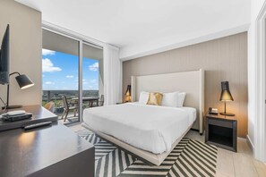 Een hotelkamer met een groot bed, een bureau met een computer en afstandsbediening, twee bedlampen en uitzicht op de stad door grote ramen.