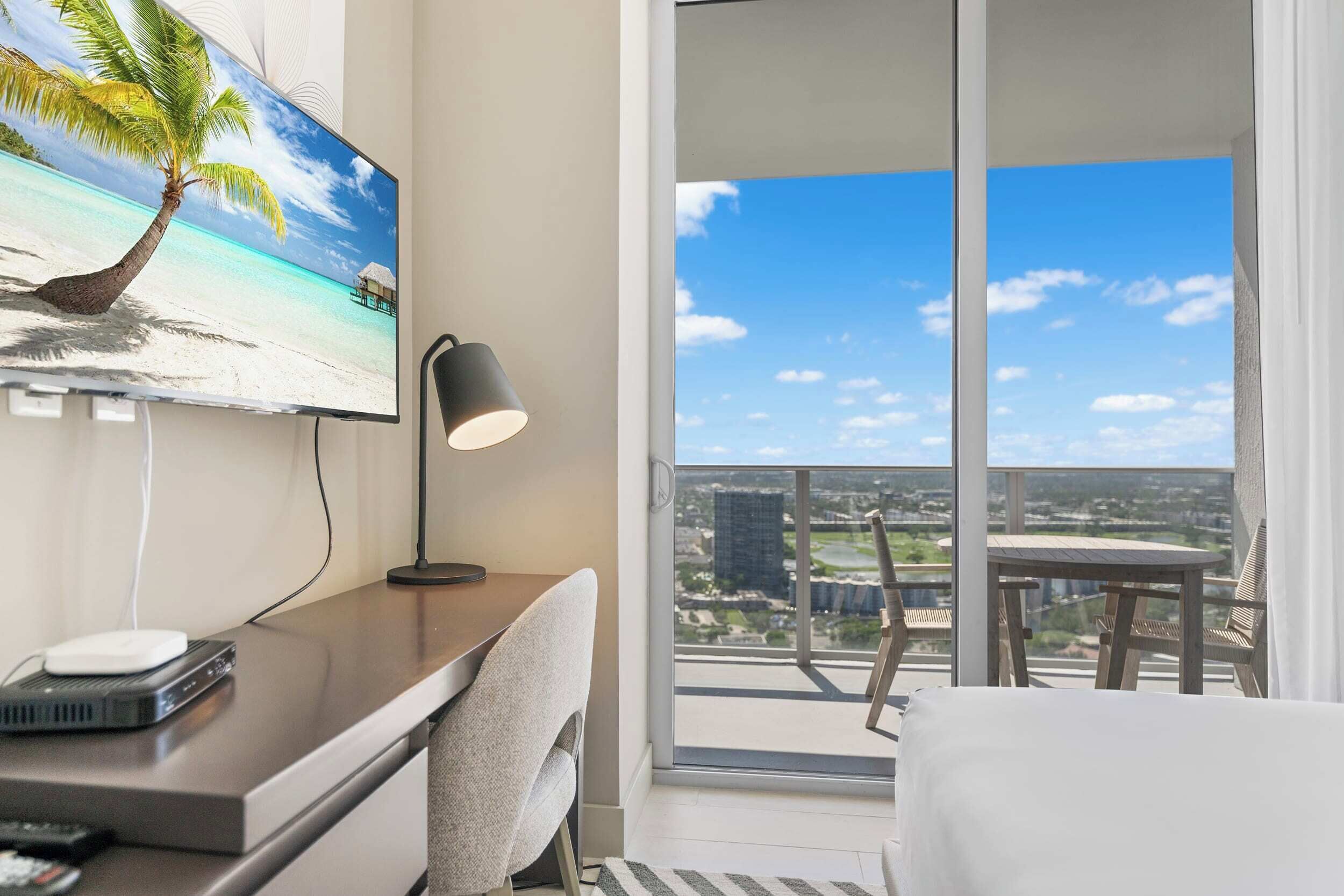 A modern hotel room with a large TV displaying a beach scene, a desk with a lamp, and a balcony with a city view.