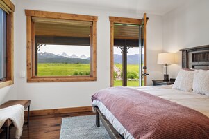 Un dormitorio con una cama grande, cabecera de madera, un banco, una lámpara y vistas a montañas a través de una ventana.