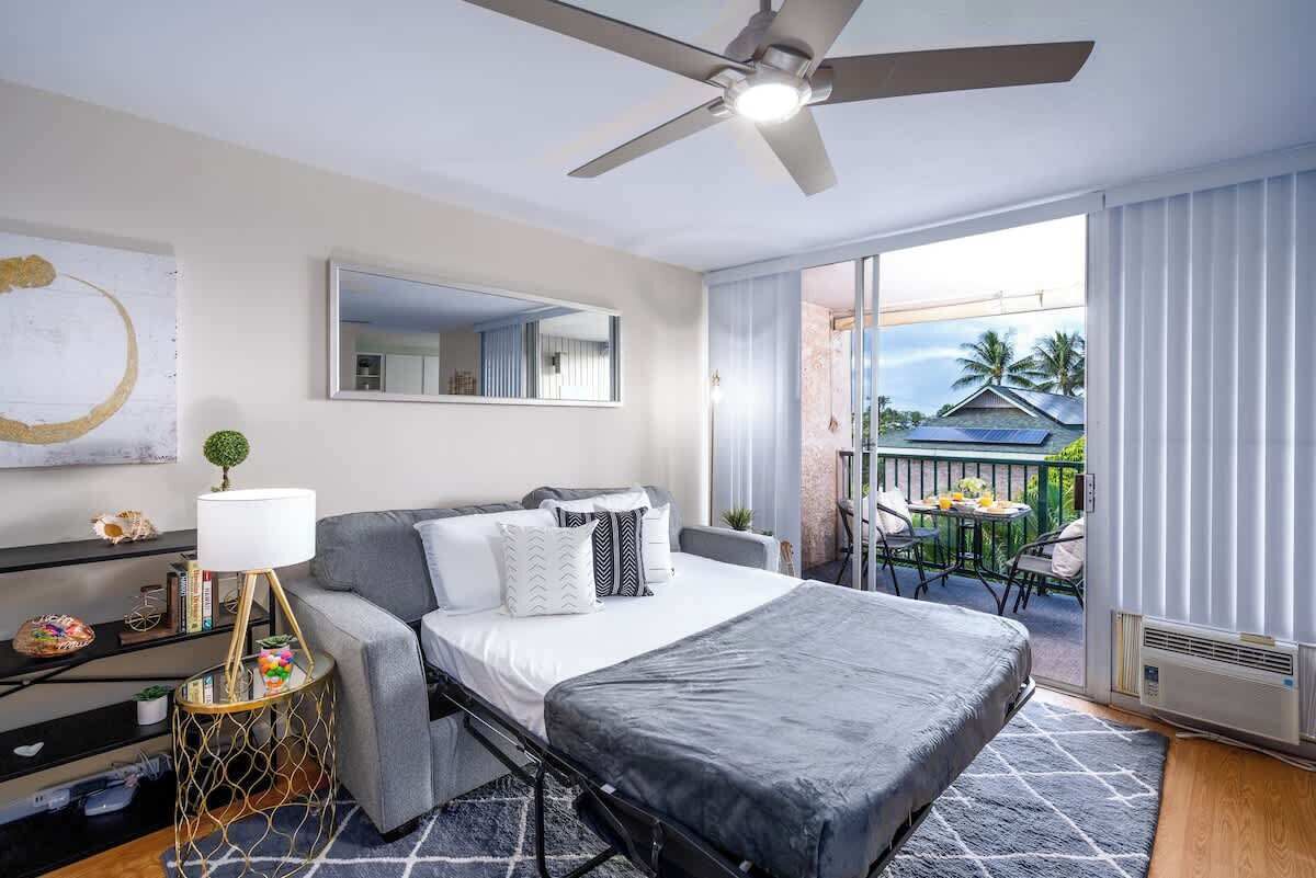 A modern hotel room with a bed, a desk, a chair, a balcony with a view of palm trees and a house.