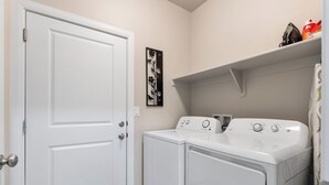 A laundry room with a white door, two washing machines, a shelf with items, and a wall-mounted artwork. - Casita Respite (Washington)