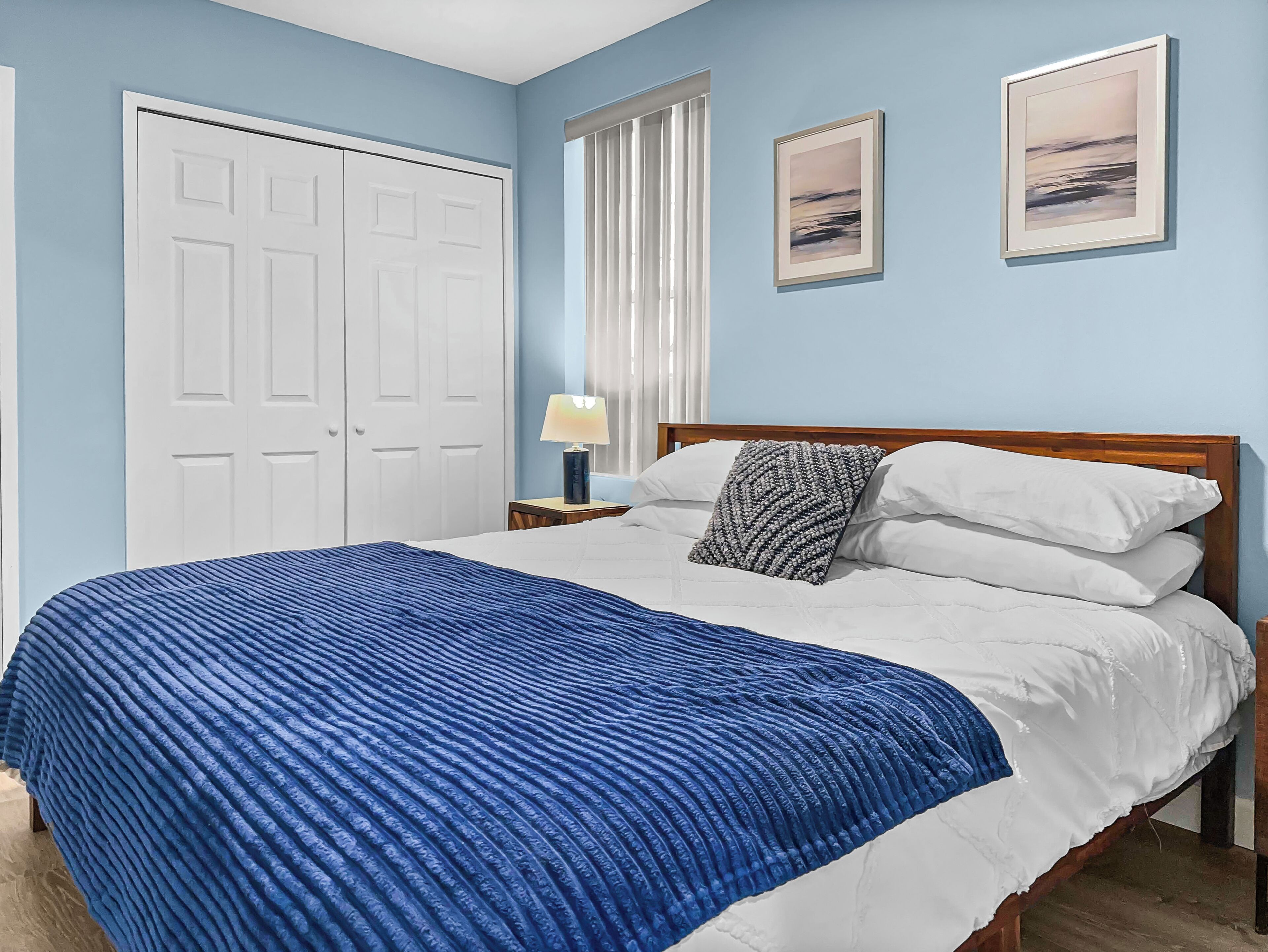 A bedroom with a bed, white pillows, a blue blanket, a wooden headboard, a nightstand with a lamp, a window with blinds, and two framed pictures on the wall.