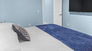 A bedroom with a bed, a blue blanket, a white pillow, a white door, and a wall-mounted TV. - Blue Haven A (Sarasota)