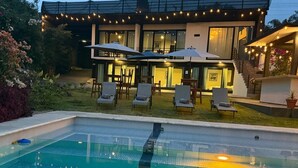 A swimming pool with lounge chairs and umbrellas, surrounded by a house with string lights. - Villa DPJ el Arco (Valle de Bravo)