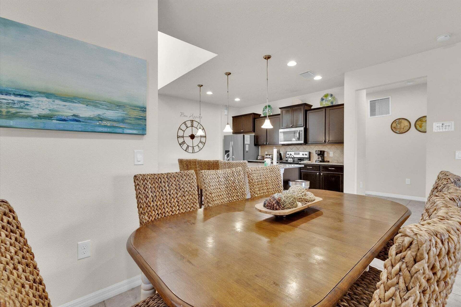 A dining area with a wooden table, woven chairs, a clock, and a kitchen in the background.