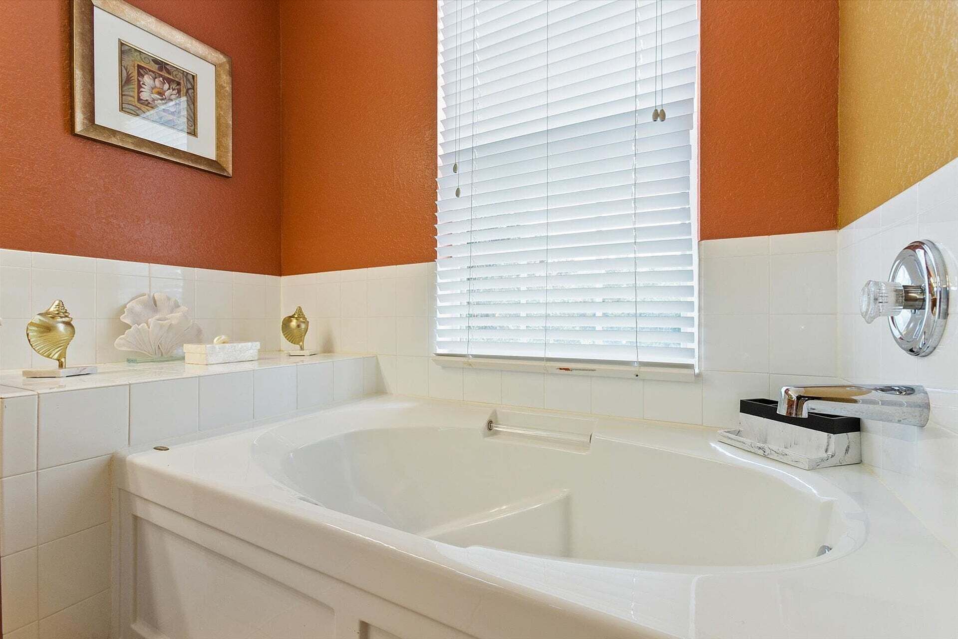 A bathroom with a bathtub, a window with blinds, and a framed picture on the wall.