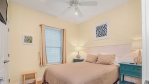 Una camera da letto con un letto, un ventilatore a soffitto, un comodino, una lampada, una finestra con tende e un piccolo sgabello.