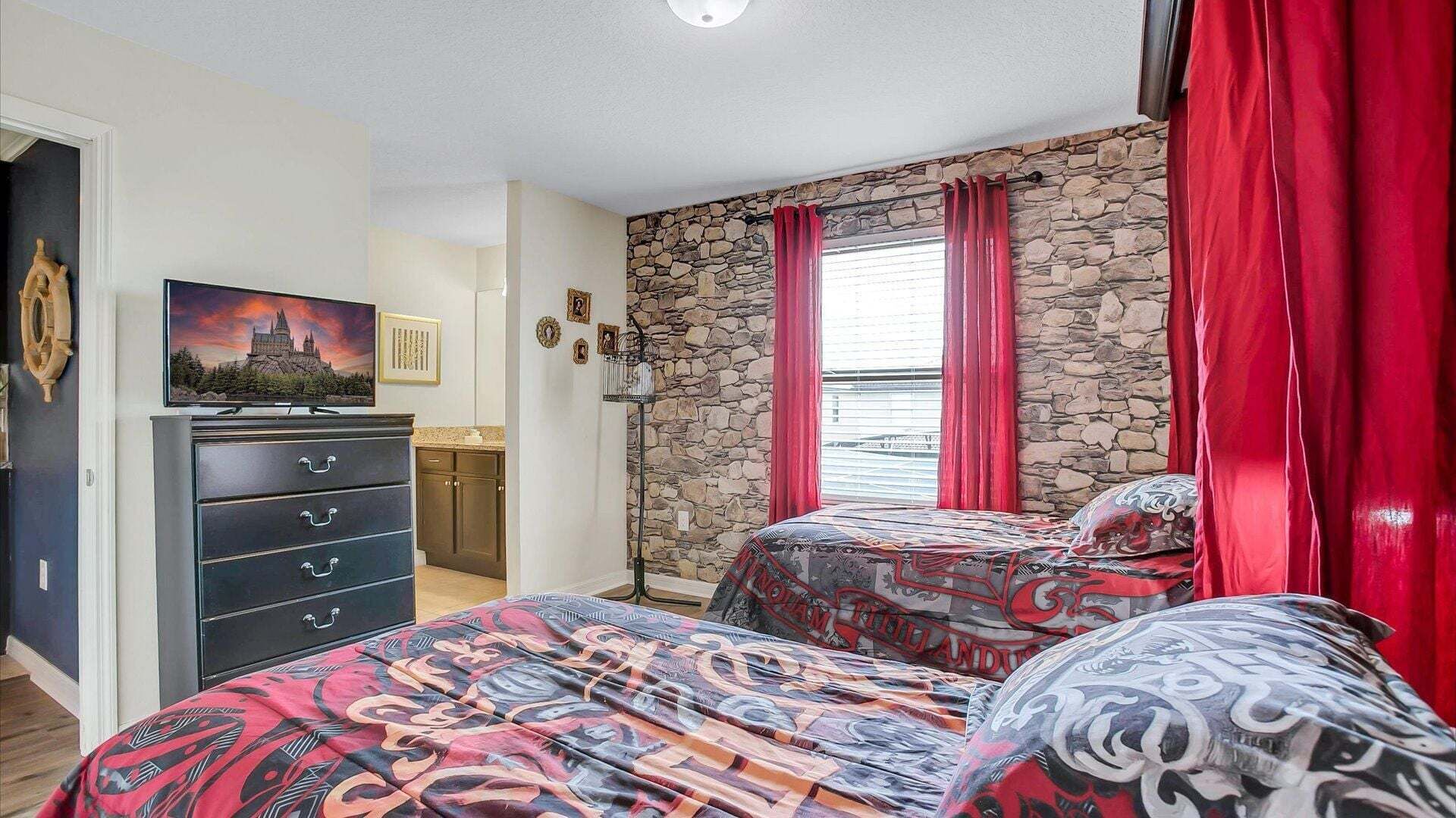 A bedroom with two beds, a dresser, a television, a stone wall, and a window with red curtains.