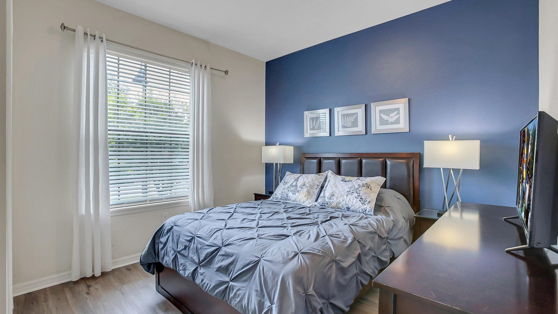 A bedroom with a bed, a nightstand, a TV, and a window with blinds.