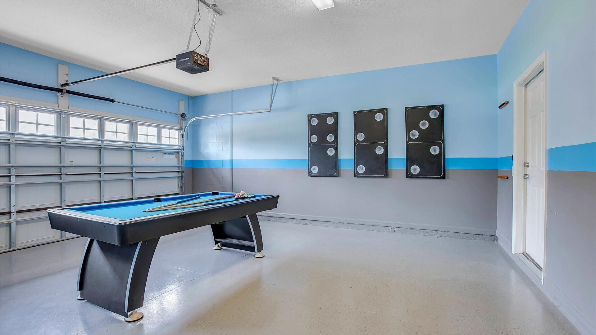 Une table de billard avec des billes, un projecteur fixé au plafond et trois haut-parleurs fixés aux murs, dans une pièce aux murs bleus et à la porte blanche.