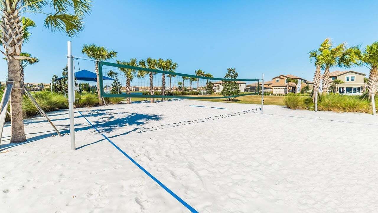 A beach volleyball court with a net, palm trees, and houses in the background.
