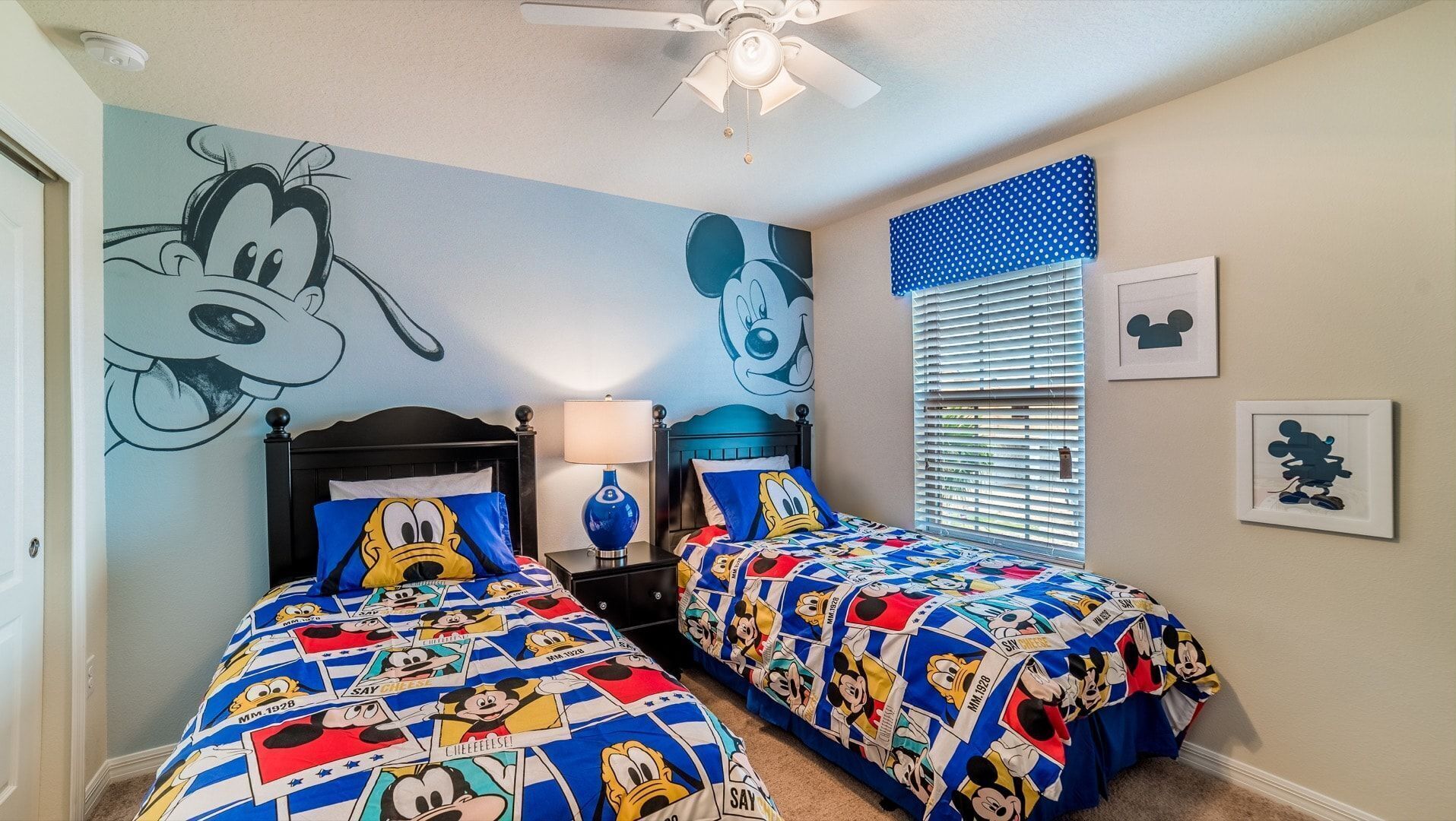 Une chambre avec deux lits, chacun agrémenté de linge de lit aux motifs de Mickey Mouse, un ventilateur de plafond et une fenêtre avec des stores.
