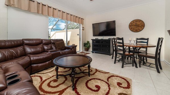 A living room with a brown leather sofa, a round coffee table, a TV, a wooden dining table with chairs, and a clock on the wall. - Windsor Hills Resort 3 Bedroom Private Pool Townhome 0874 (Kissimmee)