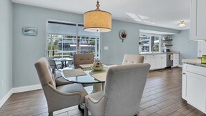 Een moderne keuken met witte kasten, een eethoek met een glazen tafel en stoelen, en een groot raam met uitzicht op de omgeving.