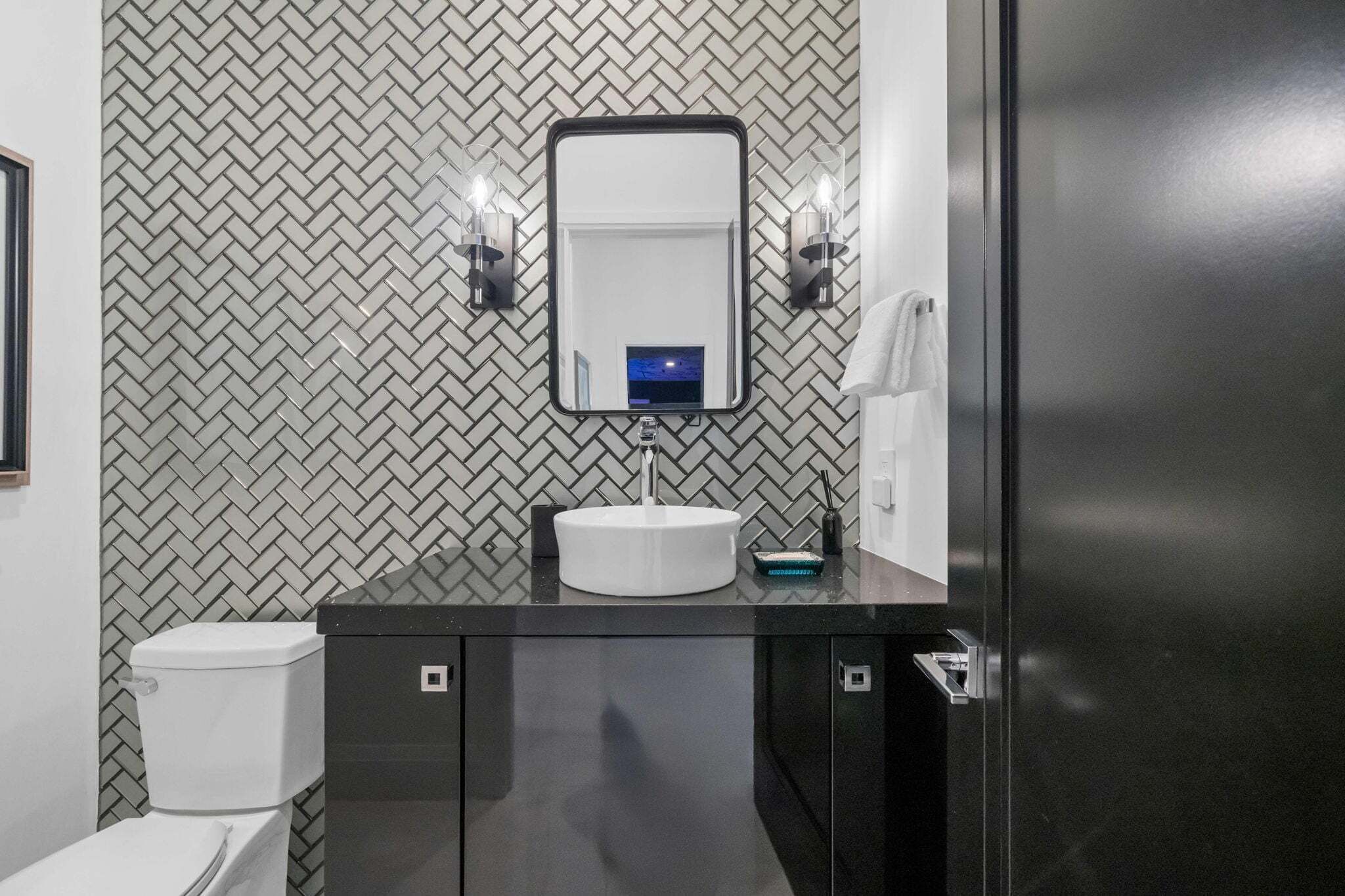 A modern bathroom with a herringbone tile backsplash, a white sink, a black vanity, and a mirror with wall-mounted lights.