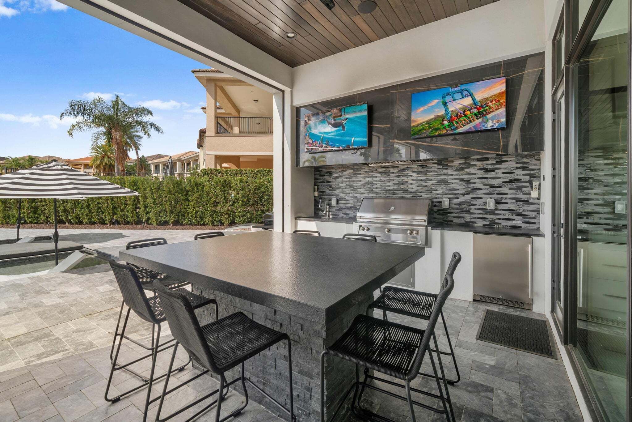 An outdoor kitchen with a stone countertop, bar stools, a grill, and a TV mounted above the counter.