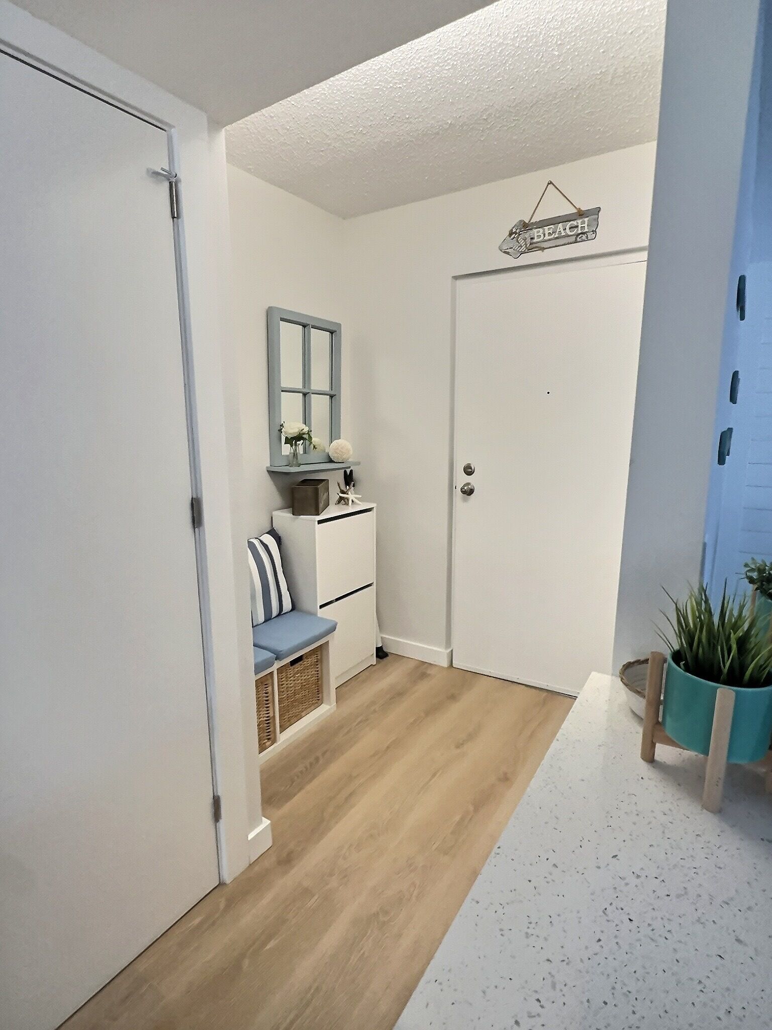 A hallway with a wooden floor, a white door, a bench with striped cushions, a small table with a vase and a ball, and a sign that reads "BEACH".