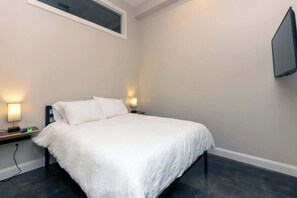 A bedroom with a bed, bedside lamps, a TV mounted on the wall, and a window. - Bright Modern Loft for Adventurers (Roanoke)