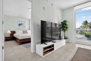 Ein modernes Schlafzimmer mit großem Fernseher, einem Bett, einem Nachttisch mit Lampe und Blick auf die Stadt durch große Fenster.