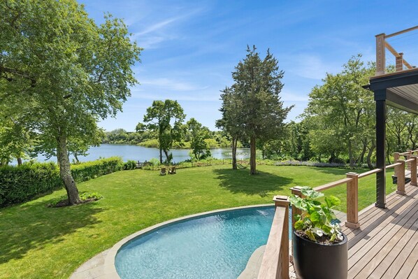A swimming pool with a wooden deck, surrounded by lush greenery and a lake view. - Kellis Cove by Rove Travel Home w Outdoor Spaces (Water Mill)