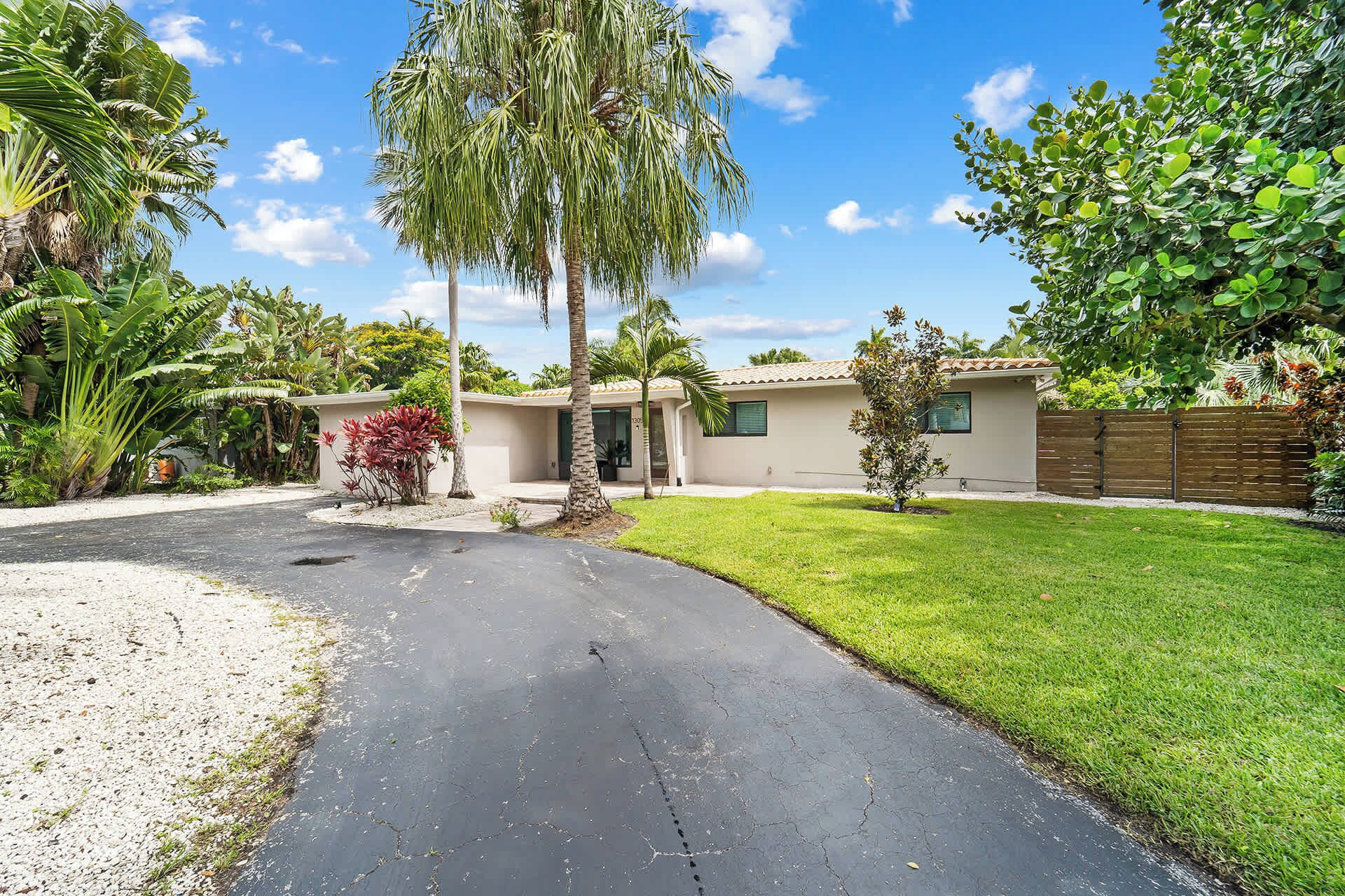 A residential property with a driveway leading to a house, surrounded by lush greenery and palm trees.