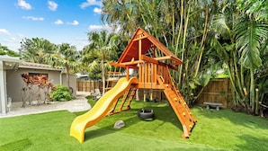 Un'area giochi con scivolo e strutture per arrampicata, circondata da un cortile recintato e palme.