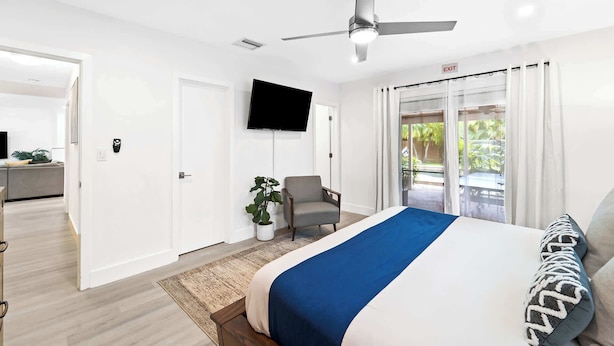 Una camera d'albergo con un letto, una sedia, una TV, una ventola a soffitto e vista sull'esterno.