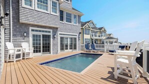 A wooden deck with a pool, white Adirondack chairs, and a two-story house with a balcony. - Dunecrest by Rovetravel - Family Oceanfront With Heated Pool (Westhampton Beach)