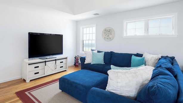A modern living room with a large flat-screen TV, a blue sofa, and a white cabinet. - Dunecrest by Rovetravel - Family Oceanfront With Heated Pool (Westhampton Beach)
