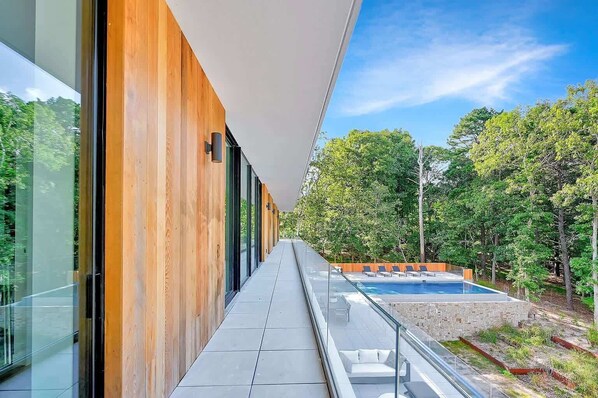 A modern building with a glass facade, a pool, and a wooden deck overlooking a forest. - Brightwater by Rove Sauna, Pool & Outdoor Spaces (East Hampton)