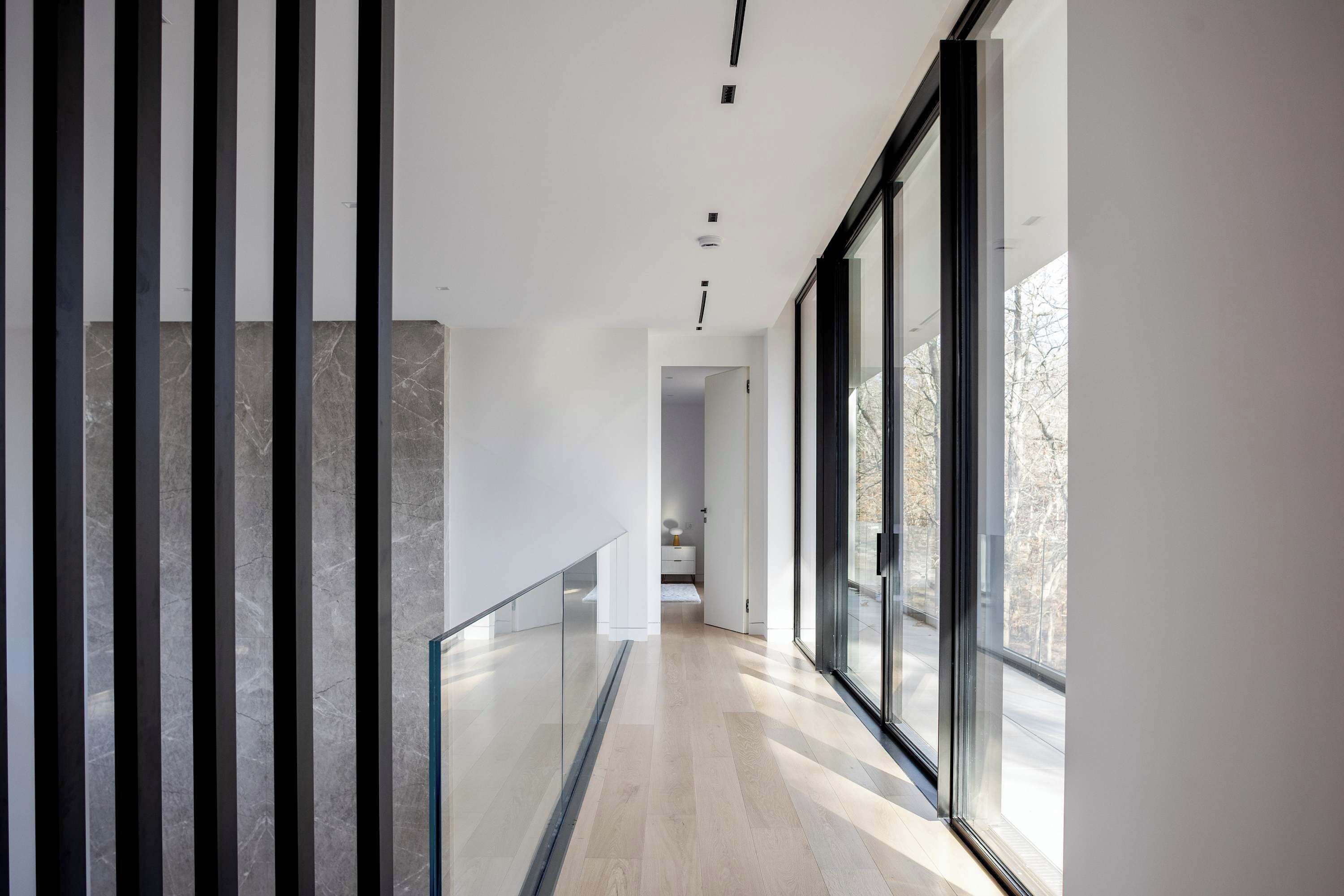 A modern hallway with a glass railing, a wooden floor, and large windows offering a view of the outside.