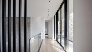 Un pasillo moderno con barandilla de vidrio, piso de madera y ventanas amplias que ofrecen vistas al exterior.