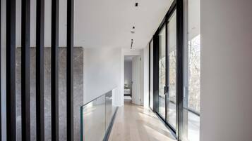 A modern hallway with a glass railing, a wooden floor, and large windows offering a view of the outside.