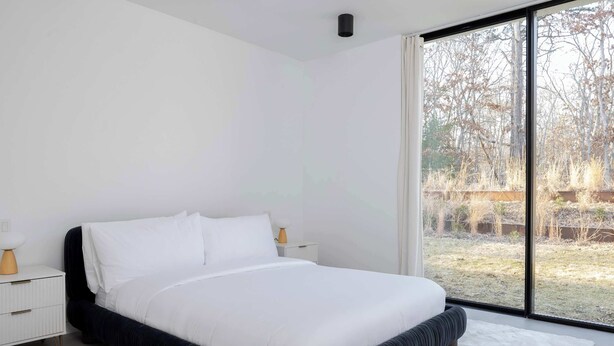 Un dormitorio moderno con una cama grande, ropa de cama blanca, una mesita de noche blanca y vistas a un jardín a través de un ventanal.