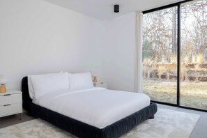 A modern bedroom with a large bed, white bedding, a white nightstand, and a view of a yard through a large window. - Brightwater by Rove Sauna, Pool & Outdoor Spaces (East Hampton)