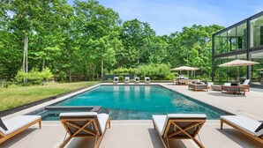 A swimming pool with lounge chairs and umbrellas, surrounded by lush greenery and modern architecture. - Glass House by Rove Travel Heated Pool & Hot Tub (East Hampton)