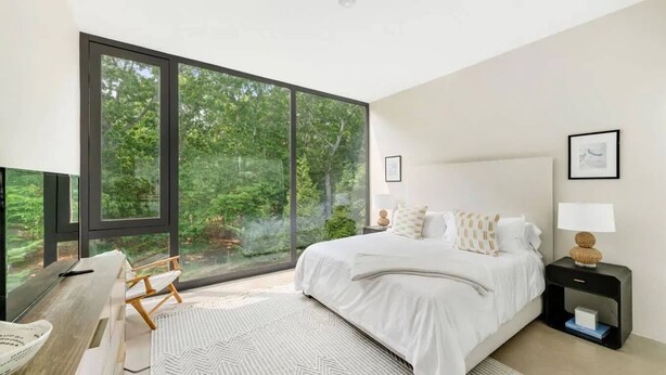 A modern bedroom with a large window, a bed, a bedside table, and a chair. - Glass House by Rove Travel Heated Pool & Hot Tub (East Hampton)