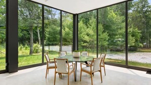 A modern dining area with a glass table and chairs, surrounded by large windows offering a view of a forested area. - Glass House by Rove Travel Heated Pool & Hot Tub (East Hampton)