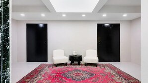 A room with a large red patterned rug, two white chairs, a small black table, and two black doors. - Glass House by Rove Travel Heated Pool & Hot Tub (East Hampton)