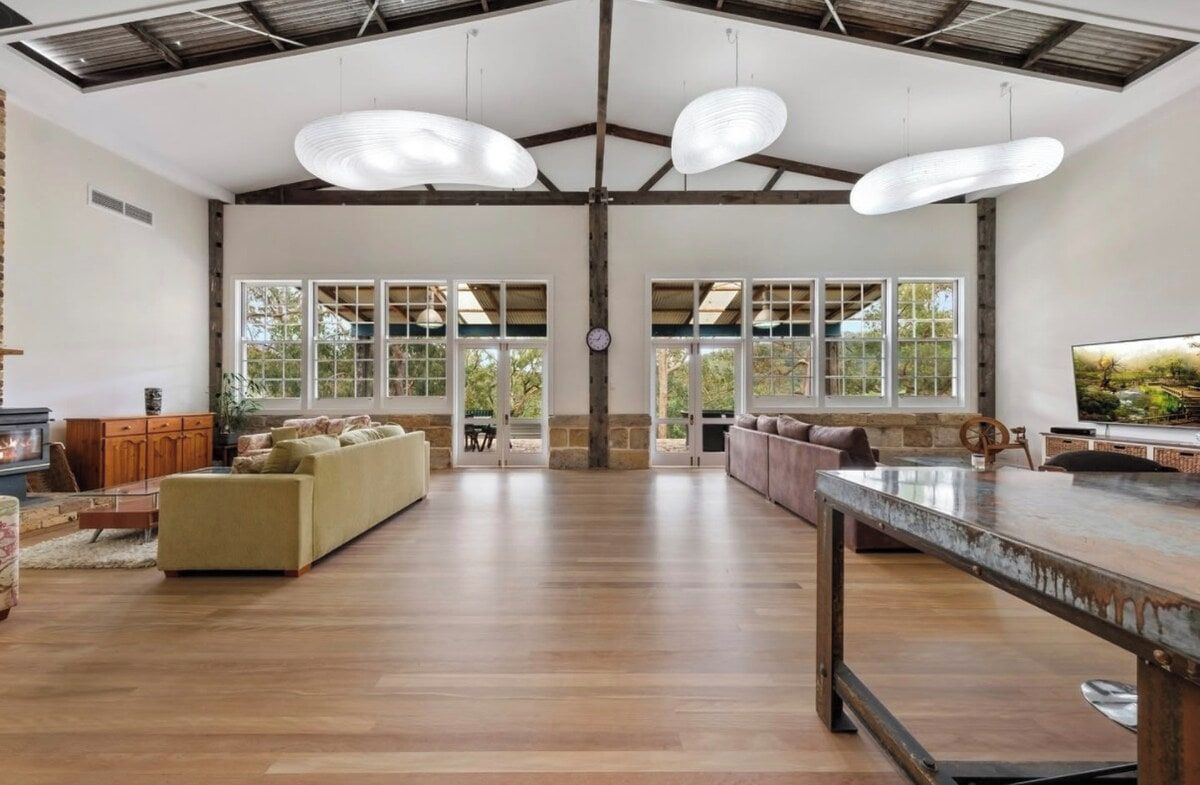 Un amplio salón con un sofá grande, una mesa de centro y un televisor sobre una mesa de madera. Hay ventanas grandes y una chimenea.