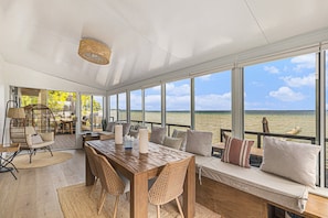 A spacious room with a large wooden table, wicker chairs, and a cushioned bench by a large window overlooking a body of water. - Waterfront Retreat on West Bay w Hot Tub, Games & More! 7345 (Traverse City)