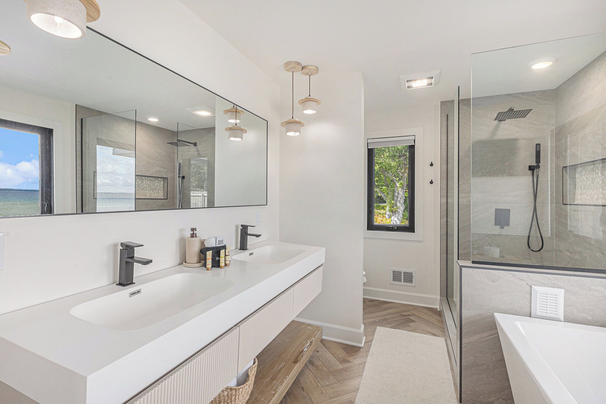 A modern bathroom with a large mirror, two sinks, a shower, and a view of the sea.